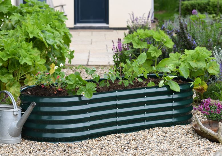 Image shows a garden where a dark-green coloured corrugated oval metal planter is placed on a grey gravel area. The planter has plants growing in its compost, including a strawberry plant in blooms with white flowers. There is a metal watering can beside the planter and borders growing flowers each side. In the blurred background is a pale stone patio and, beyond that, a navy blue door to a house with white walls.