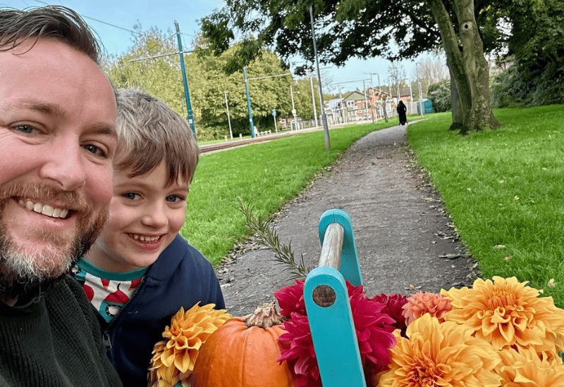 Derek from @fiveminutegardener and his son, holding a basket of flowers in a park.