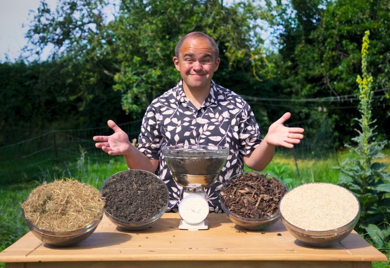 Alex Smith, stood at a table in a sunny garden, with 5 bowls of compost covered in different types of mulch.