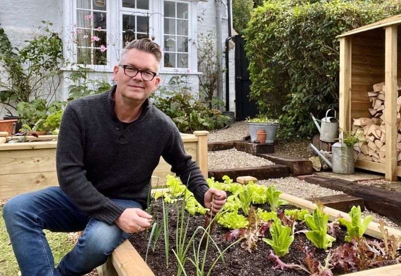A gardener sitting on the edge of a raised bed using a hand tool to cultivate soil around fresh vegetables outside a white cottage.