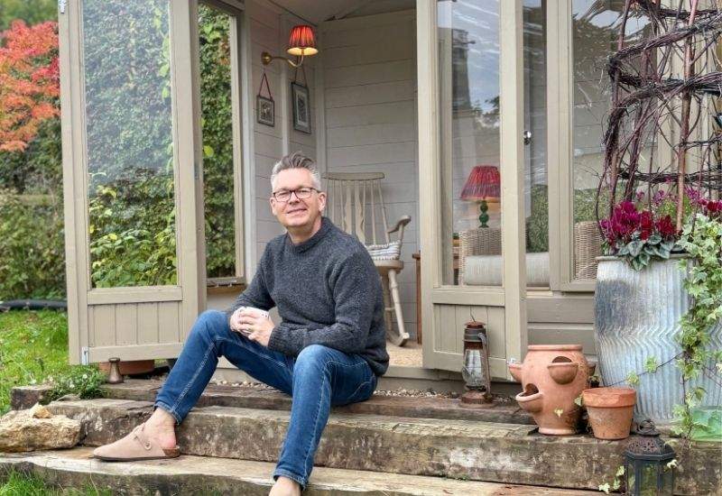 Man in a grey sweatshirt sitting on the steps of a garden summerhouse holding a mug.