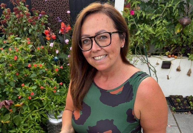 Smiling woman with brown hair and glasses wearing a green patterned top in a lush garden setting.