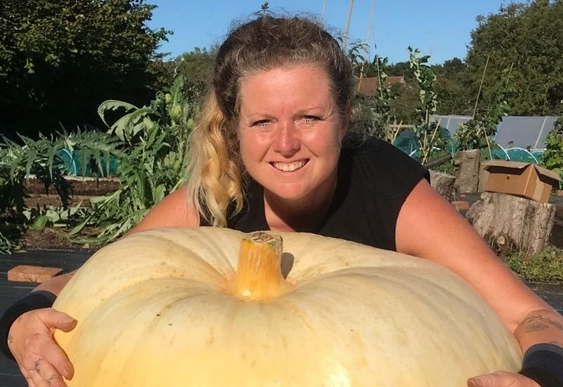 Smiling gardener hugging a giant pumpkin in an allotment garden.
