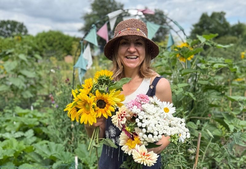 A smiling woman wearing a sun hat and overalls, holding a large bouquet of sunflowers, dahlias, and daisies in an allotment.