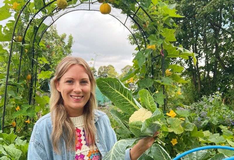 Woman in denim jacket holding freshly harvested cauliflower in garden with metal archway trellis.