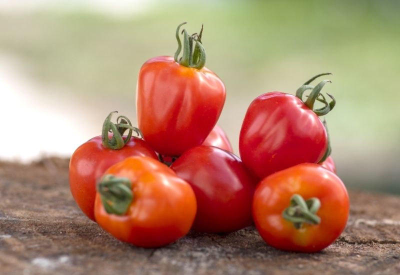 Fresh red heart-shaped tomatoes (Tomate Fraise) stacked on a wooden surface with a blurred green background.