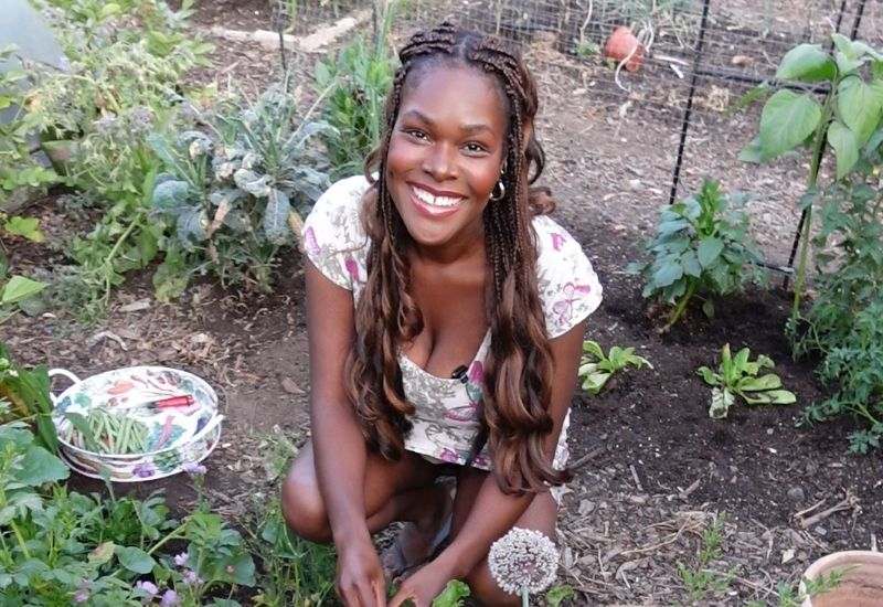 Woman smiling in vegetable garden allotment, holding a bowl of freshly harvested okra surrounded by leafy greens.