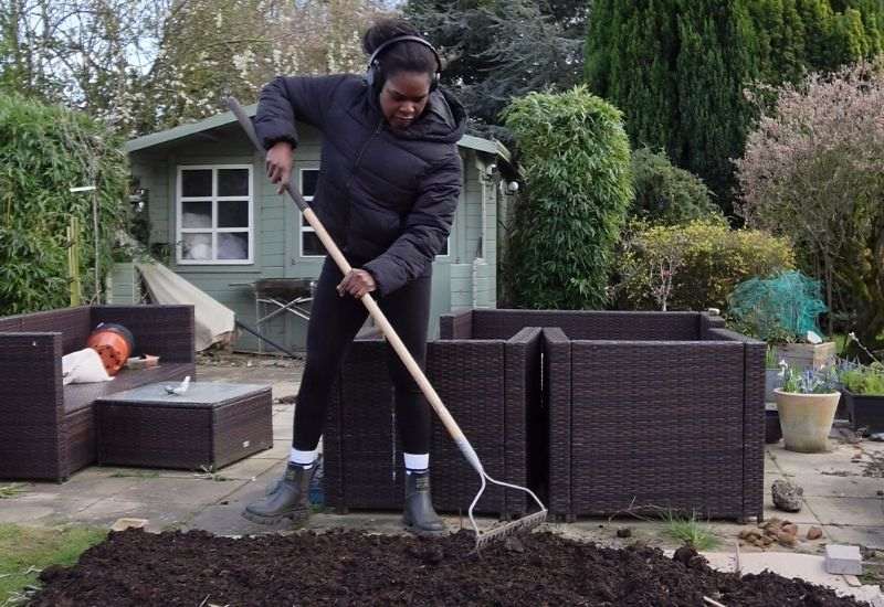 Woman raking soil in a garden bed near a patio, with a green summer house and wicker planters in the background.