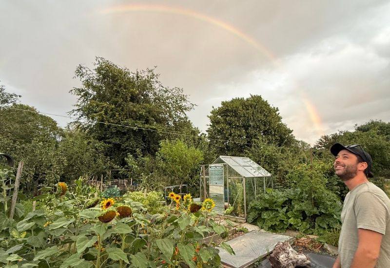 Man smiling at a rainbow over an allotment garden with sunflowers and a greenhouse.